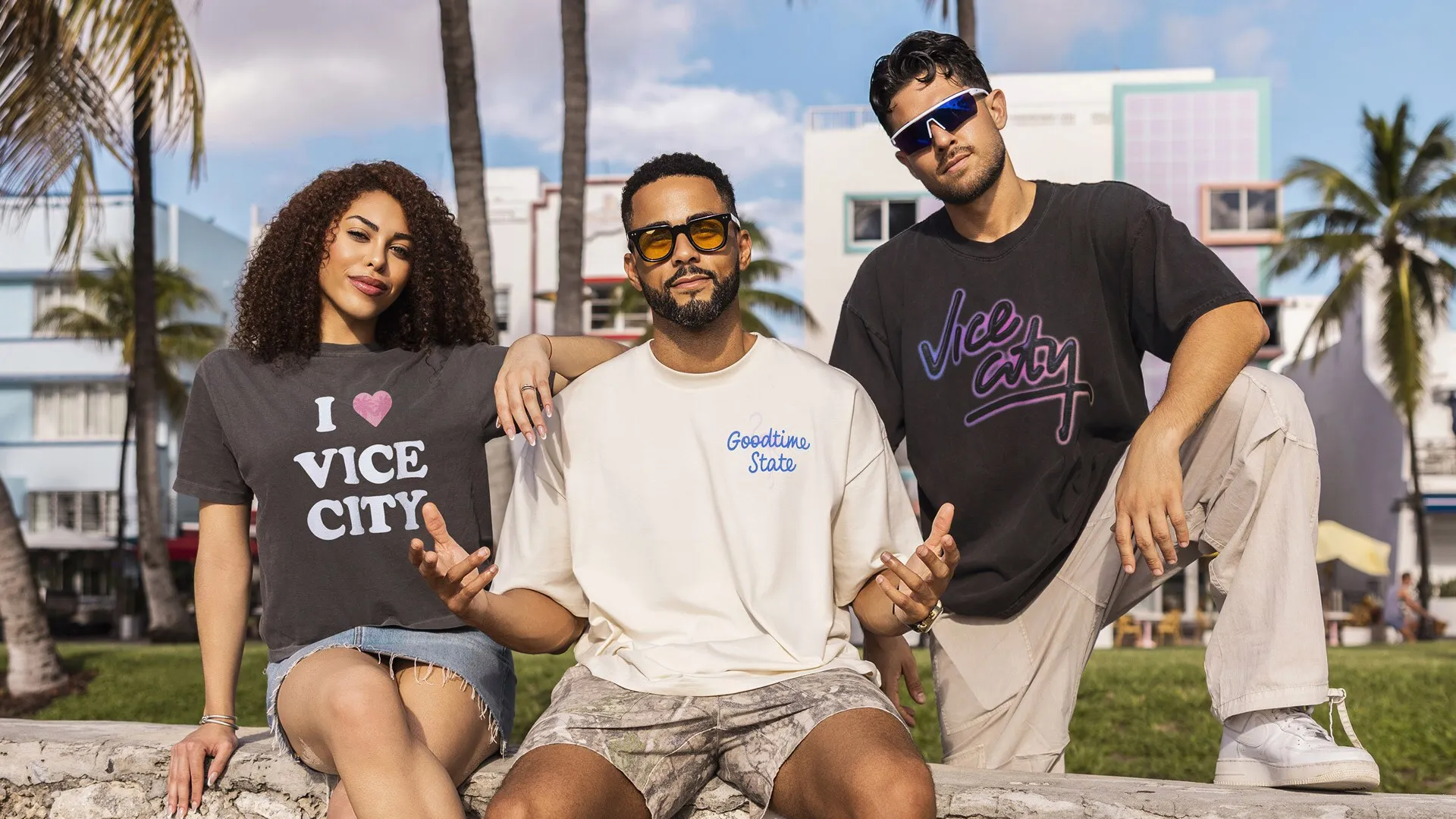 Tres personas sentadas en un muro bajo entre palmeras en Miami, con camisetas de I love Vice City, Goodtime State y Vice City.