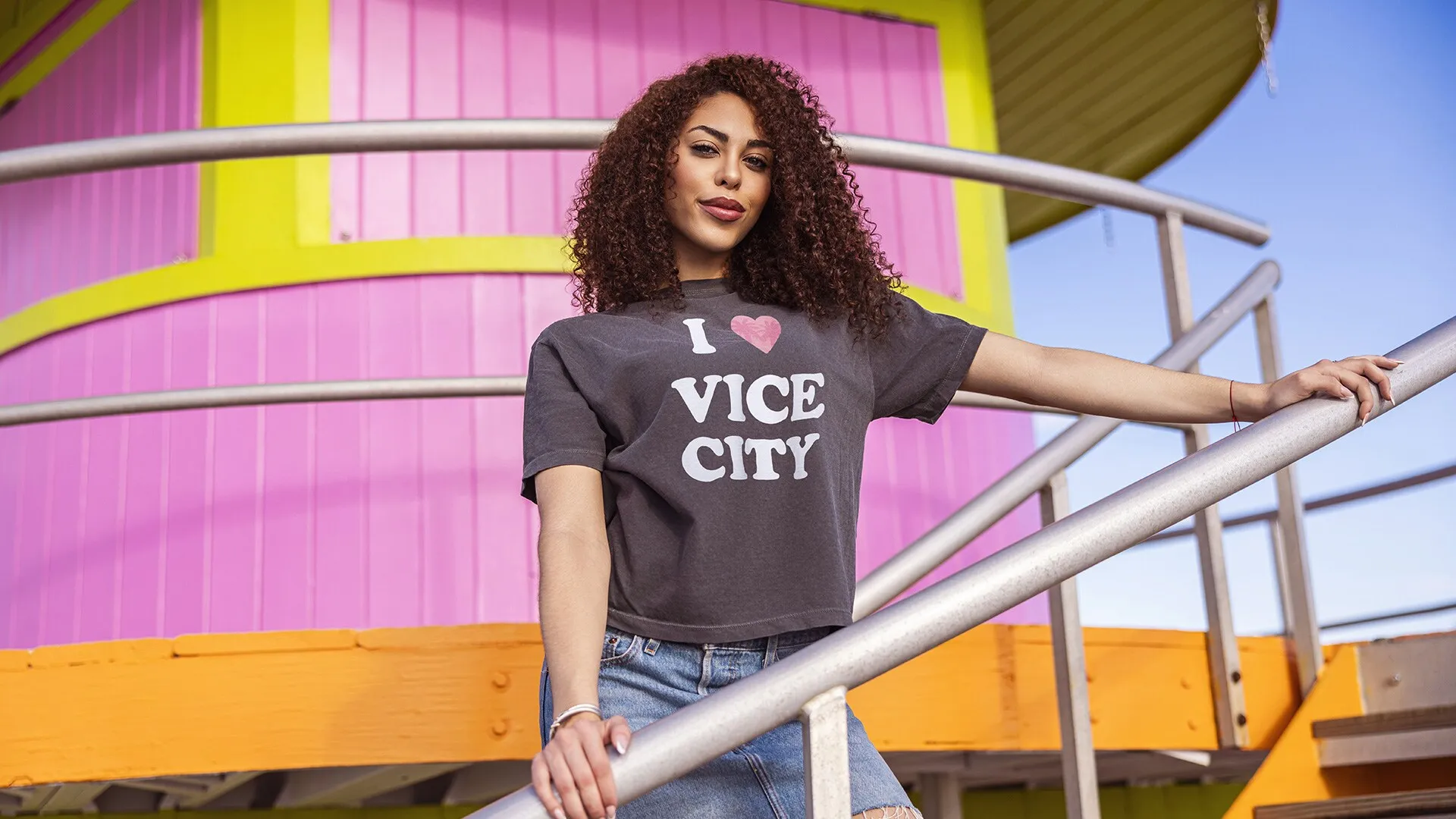 Una mujer en la escalera de una torre de socorristas rosa y amarilla, con la camiseta corta oscura I love Vice City.
