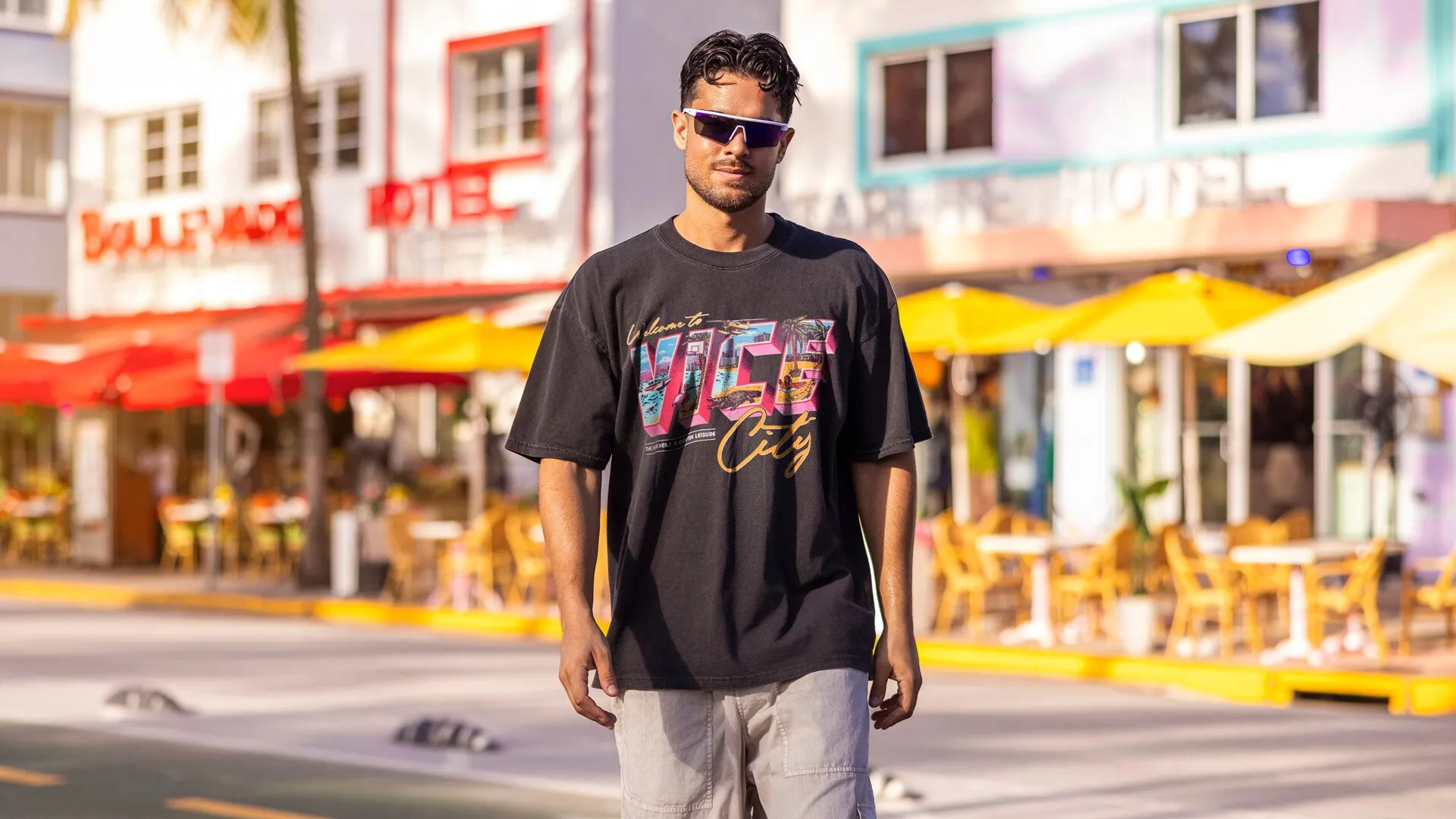 Un hombre con gafas de sol en una calle de hoteles art déco con sombrillas amarillas, con una camiseta negra con un estampado colorido de Vice City.