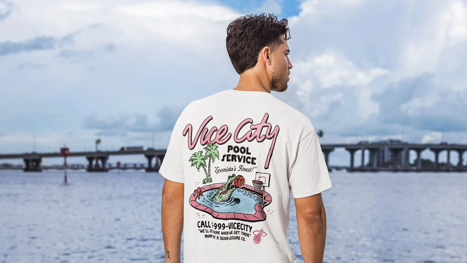 Un hombre de espaldas junto al agua con un puente a lo lejos, con la camiseta blanca Vice City Pool Service.