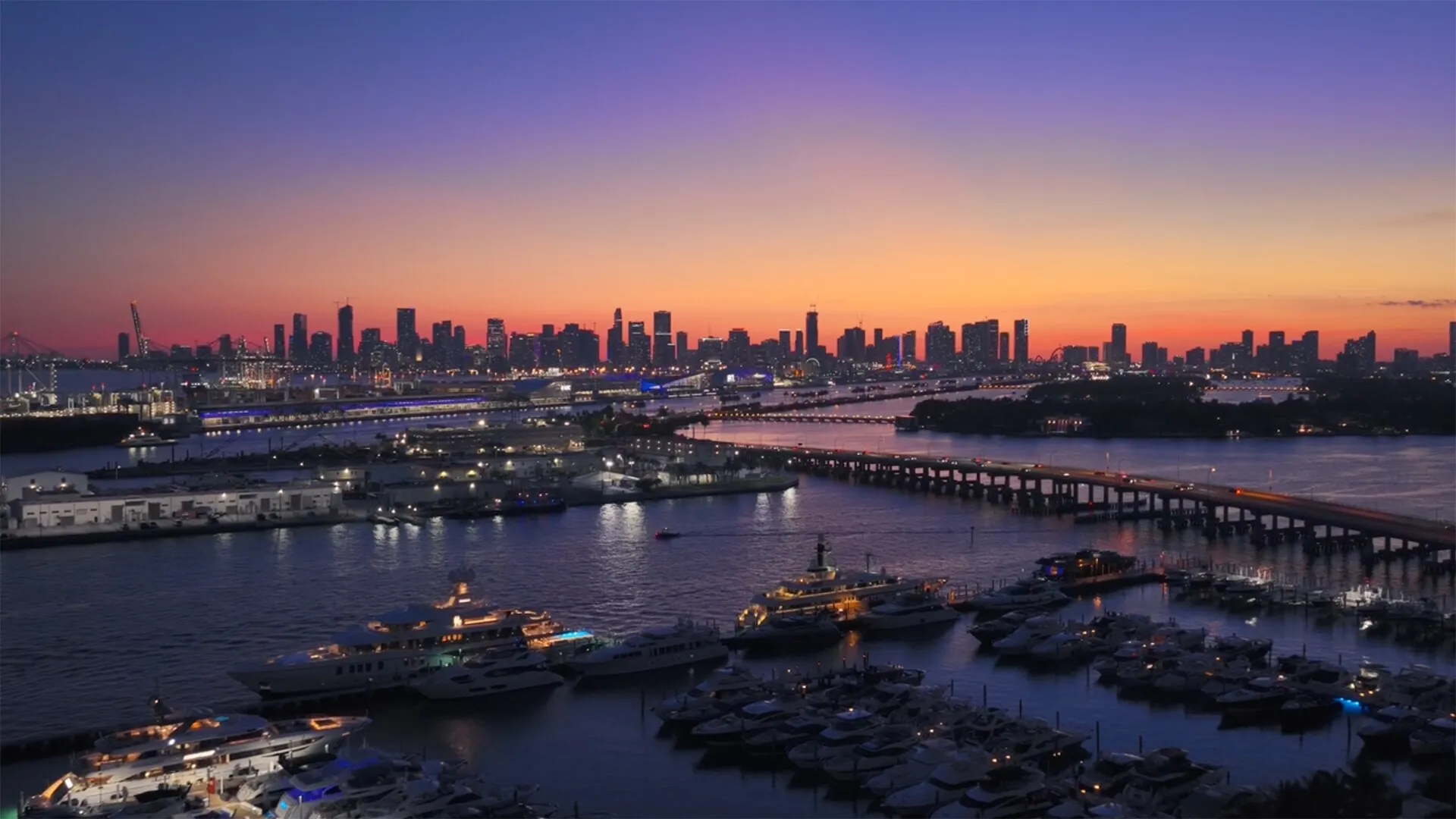 El horizonte de Miami al atardecer bajo un cielo naranja y morado, con yates en un puerto deportivo y una calzada sobre el agua.