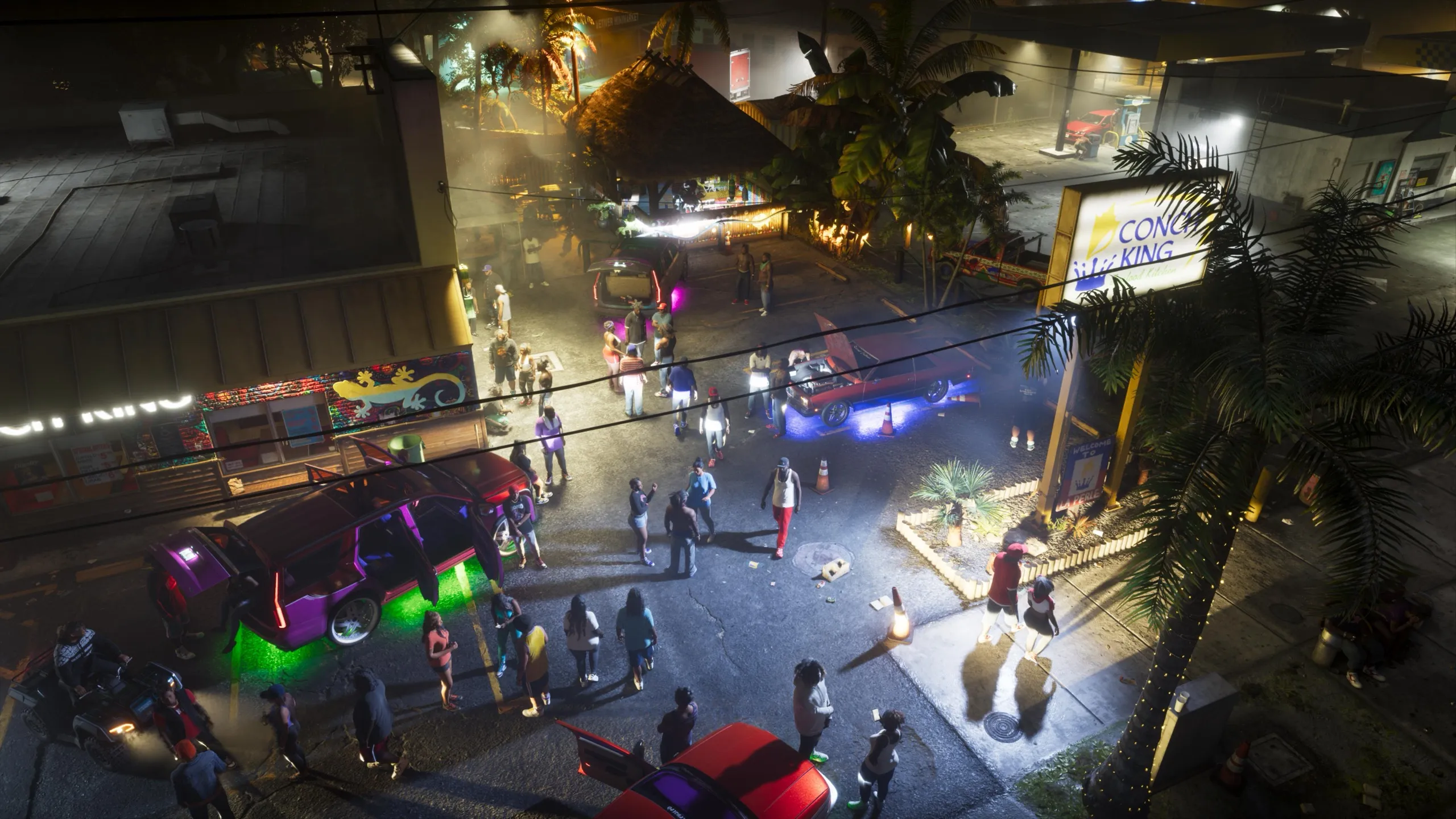 A car meet at night seen from above, with a crowd between cars lit in purple and green, next to the illuminated sign of Conch King Seafood Kitchen.
